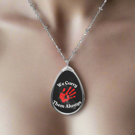 MMIW - Black pendant necklace on a silver chain. Grey smoke background with a red handprint surrounded by white lettering "We Carry them always" in honor of Missing and Murdered Indigenous Women and People. MMIW MMIP - woman's chest