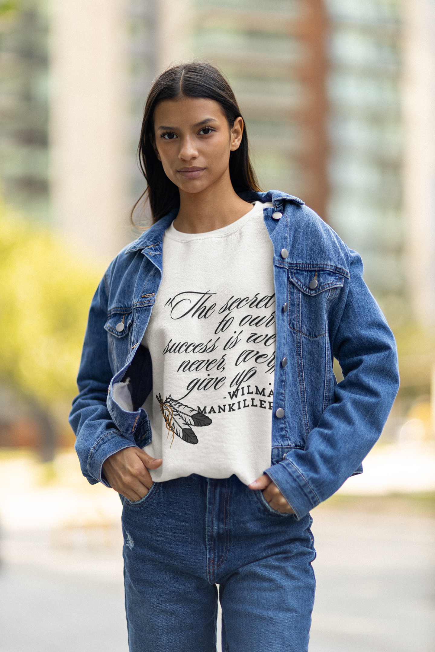 Native American Success Tshirt - Indigenous model wearing a white shirt quoting Wilma Mankiller stating the secret to our success is that we never ever give up. Eagle feathers in the corner tied together with leather string.