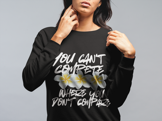 A comfy, cozy crewneck sweatshirt with the quote you can't compete where you don't compare. Three white hawaiian flowers with yellow centers lined through the middle. - black